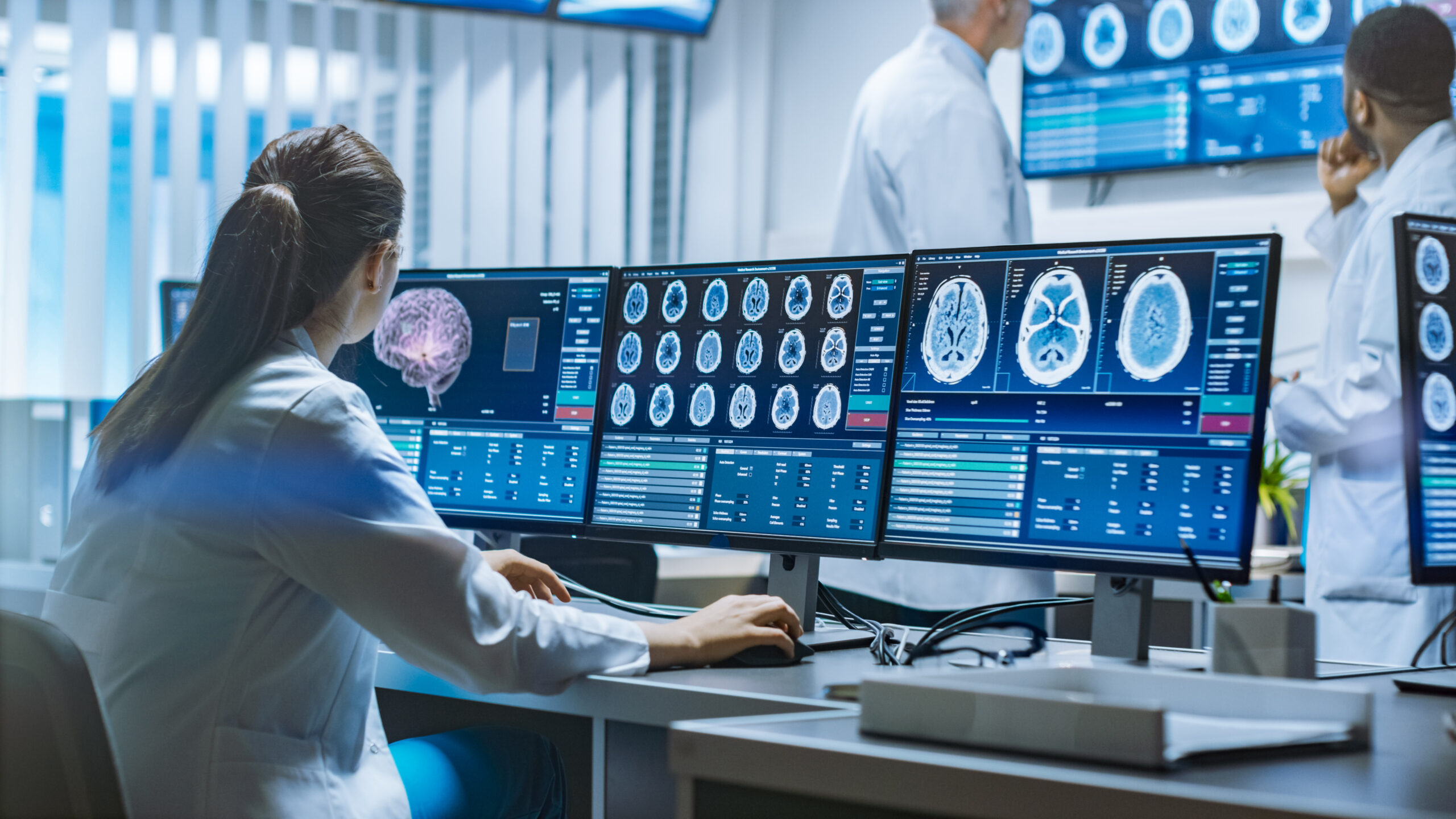 Des médecins analysent des scans cérébraux sur plusieurs écrans d'ordinateur dans une salle de contrôle d'imagerie médicale.